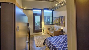 A bedroom with a bed, a sofa, a small table with a lamp, a refrigerator, and a window with a view. - Chic Ultramodern Minimalist Studio Loft + Patio (Saint Louis)