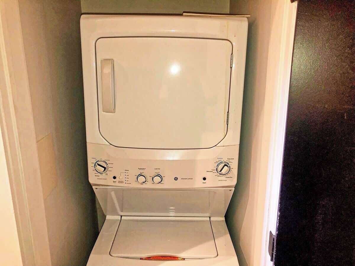Lavadora blanca compacta en un espacio reducido, con un panel de control que incluye varios diales y botones.