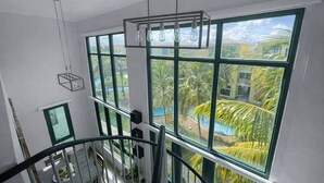 Een kamer met grote ramen, een hanglamp en uitzicht op een zwembad en palmbomen.