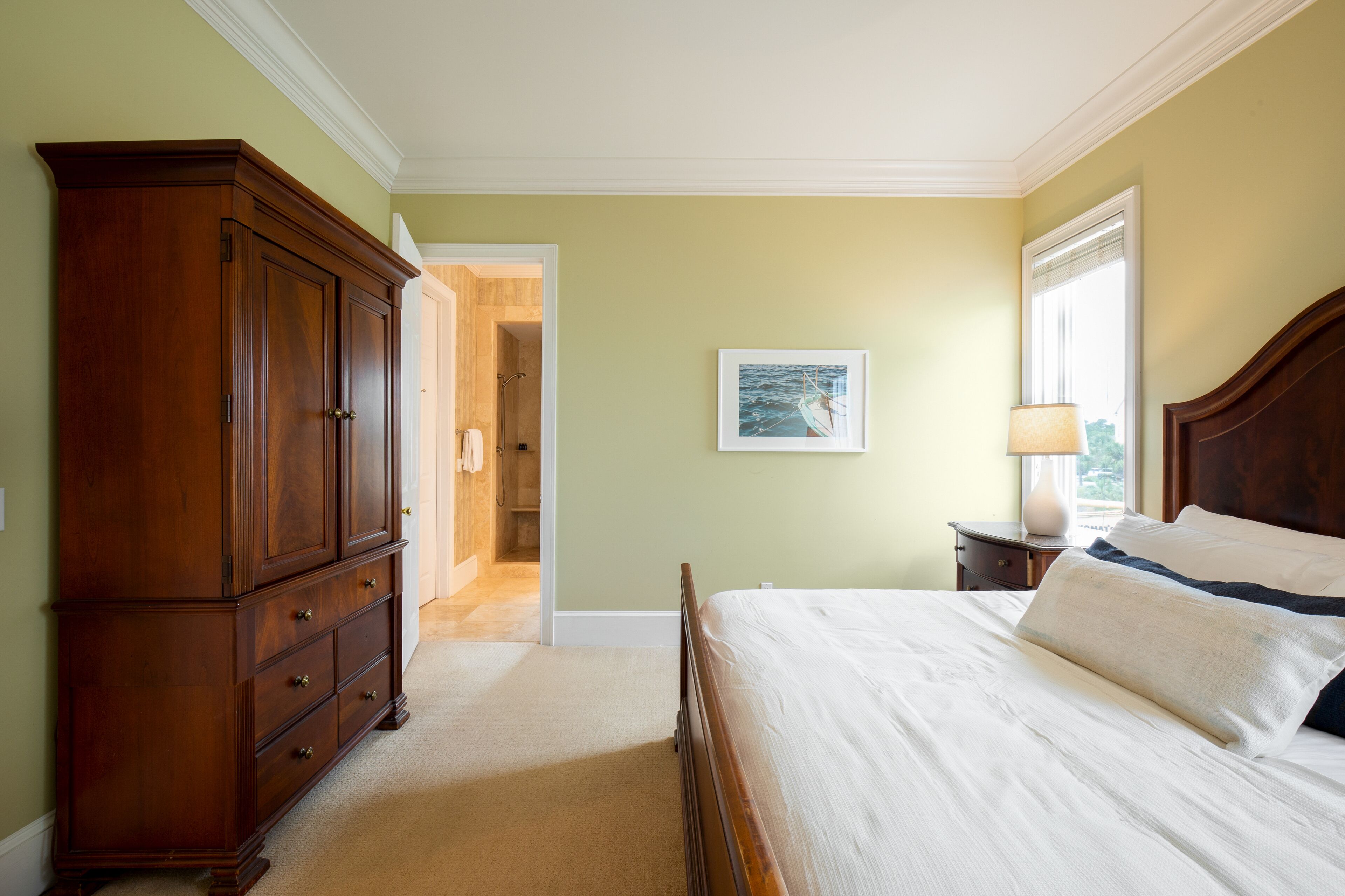 Une chambre avec une grande commode en bois, un lit recouvert de draps blancs, un tableau encadré au mur et une fenêtre laissant entrer la lumière naturelle.