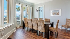 Un coin repas avec une table en bois foncé et des chaises en osier, de grandes fenêtres avec des rideaux et un lustre.
