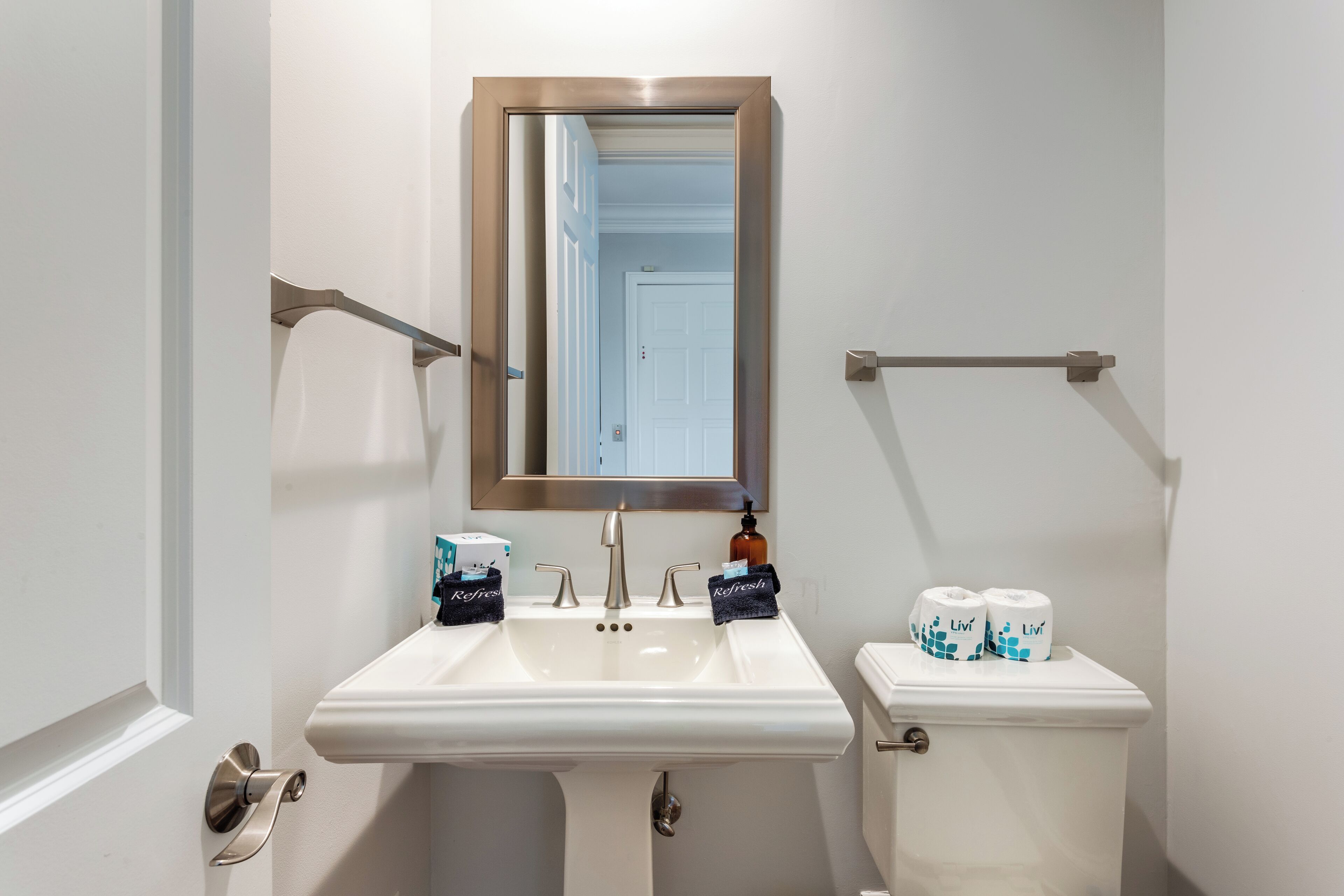 Une salle de bain avec un lavabo blanc, un miroir et des toilettes. Il y a deux rouleaux de papier toilette et un flacon de savon sur le réservoir des toilettes.