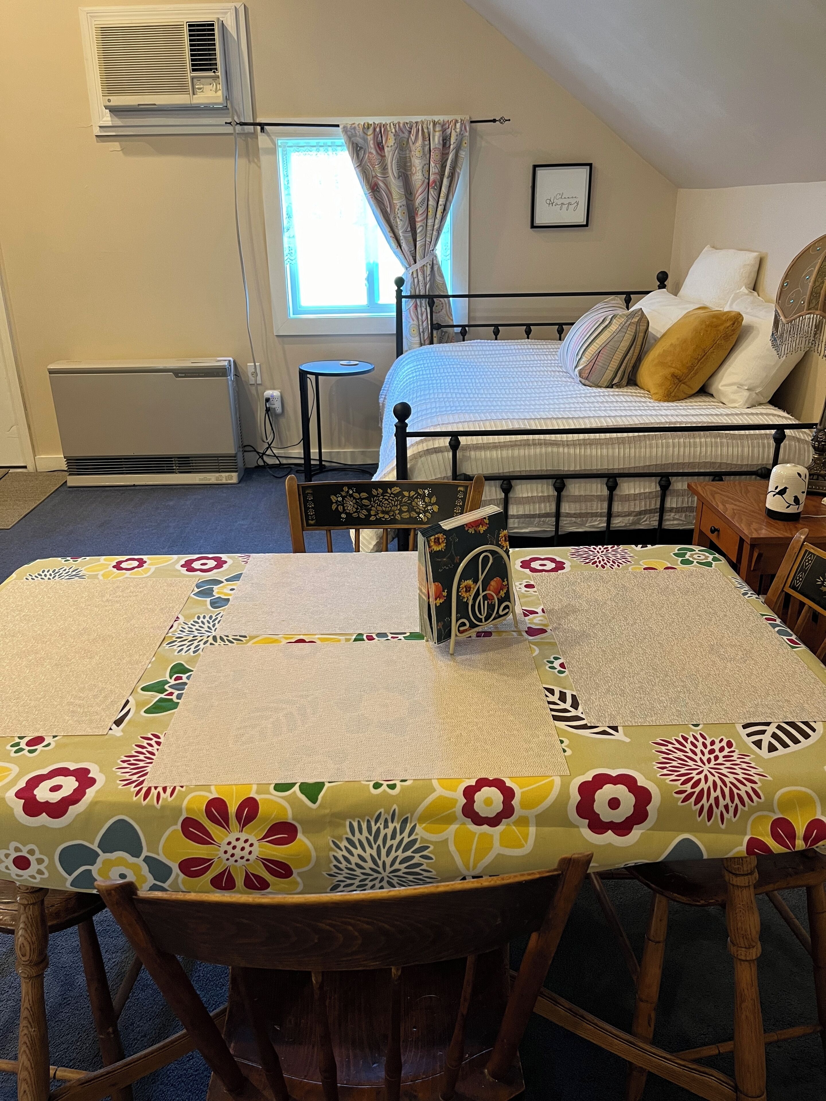 Een kamer met een bed, een tafel met een tafelkleed met bloemen, een stoel, een bijzettafeltje en een luchtverkoeler aan de muur.