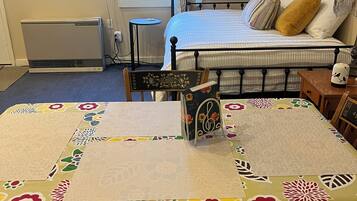 Een kamer met een bed, een tafel met een tafelkleed met bloemen, een stoel, een bijzettafeltje en een luchtverkoeler aan de muur.