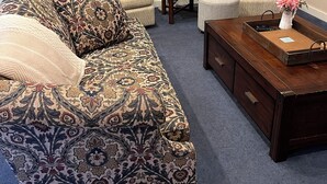 Uma sala de estar acolhedora com um sofá estampado, duas poltronas, uma mesa de centro de madeira e um aparador. Um espelho decorativo está pendurado na parede, e uma mesinha lateral fica posicionada entre as poltronas.