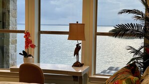 A bedroom with a large window overlooking a body of water, a bedside table with a lamp and a decorative bird statue, and a bed with a floral pillow.