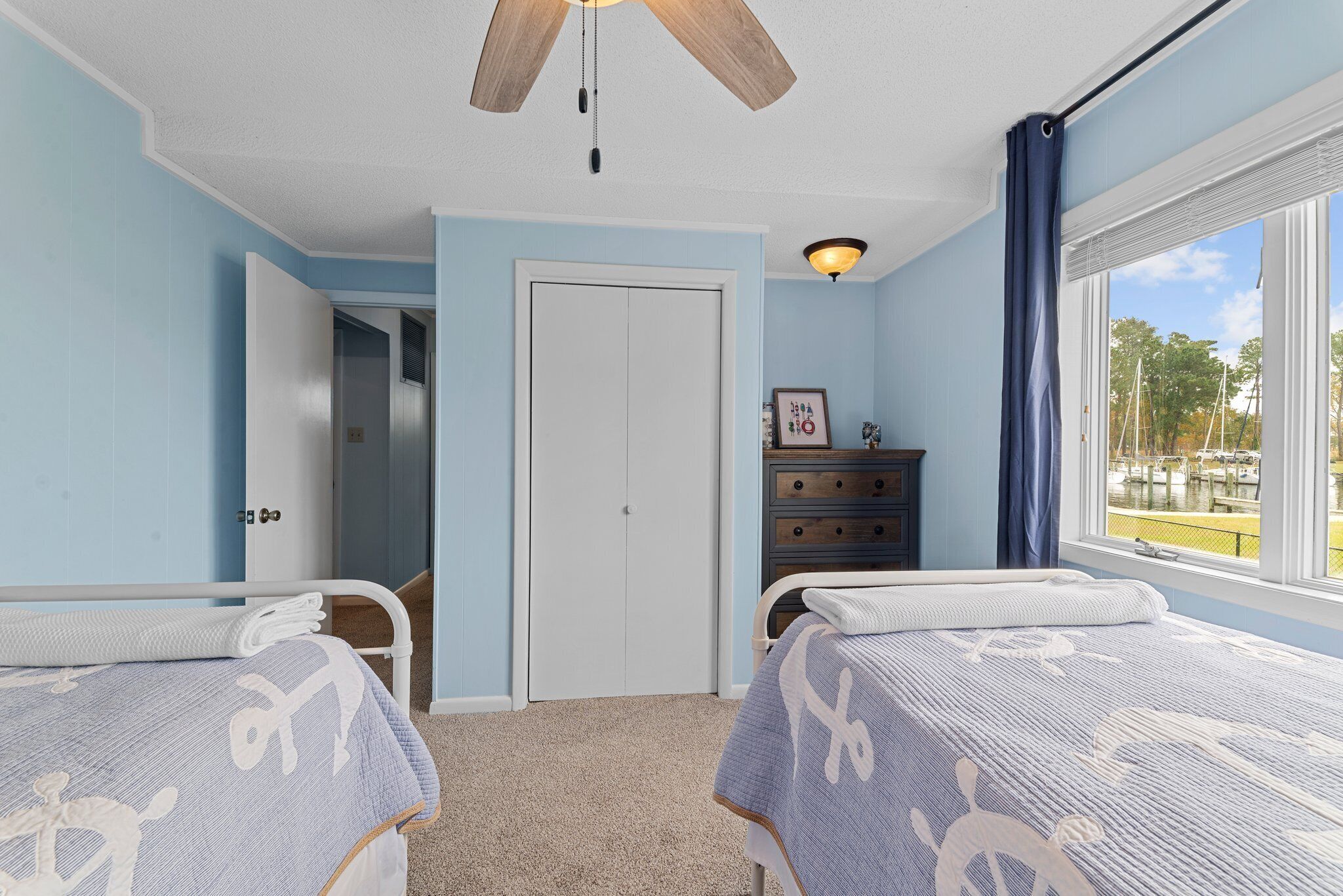 Une chambre avec deux lits, un ventilateur de plafond, une commode, une fenêtre donnant sur le port et une porte menant à une autre pièce.