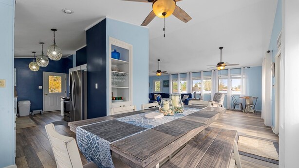 A spacious dining area with a long wooden table, white chairs, and a ceiling fan.