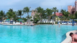 A pool area with lounge chairs and umbrellas, palm trees, and a building in the background. - Spend your holiday in Paradise (Paradise Island)
