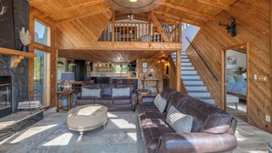 A spacious loft-style living area with a high ceiling, wooden beams, a fireplace, leather sofas, and a staircase leading to an upper level.
