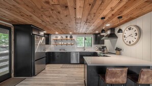 A modern kitchen with a wooden ceiling, black cabinets, a central island, pendant lights, and a large wall clock.