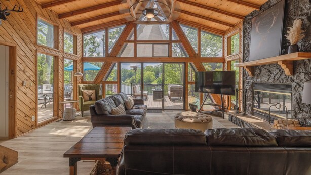 A spacious living room with a high wooden ceiling, large windows, a fireplace, and a television.