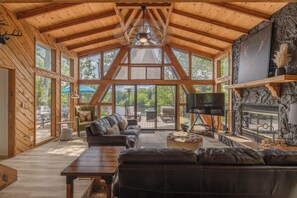 A spacious living room with a high wooden ceiling, large windows, a fireplace, and a television.