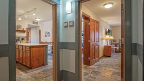 Un couloir avec un coin cuisine, des meubles de rangement en bois, un sol carrelé et une porte donnant sur une autre pièce.