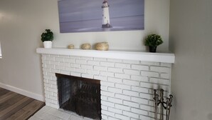 Una chimenea de ladrillo blanco con un repisa que exhibe un cuadro de un faro, jarrones decorativos y pequeñas macetas.