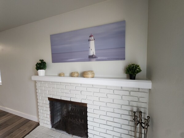 Una chimenea de ladrillo blanco con un repisa que exhibe un cuadro de un faro, jarrones decorativos y pequeñas macetas.