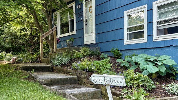 Une maison bleue avec une pancarte indiquant «Entrée des invités», un escalier en pierre et un jardin où poussent différentes espèces de plantes.