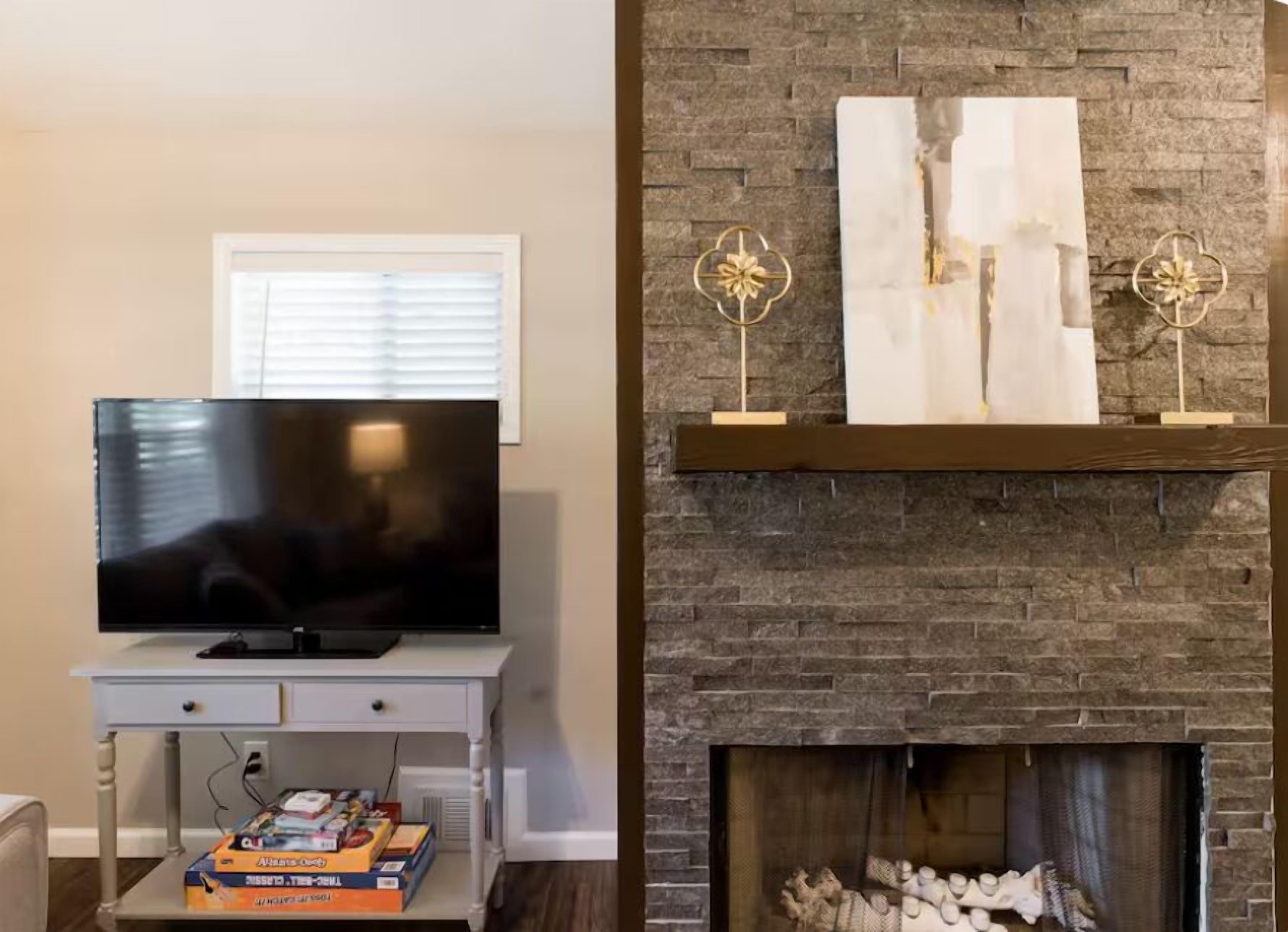A living room with a flat-screen TV on a white stand, a fireplace with a stone mantle, and two decorative gold-framed wall art pieces.