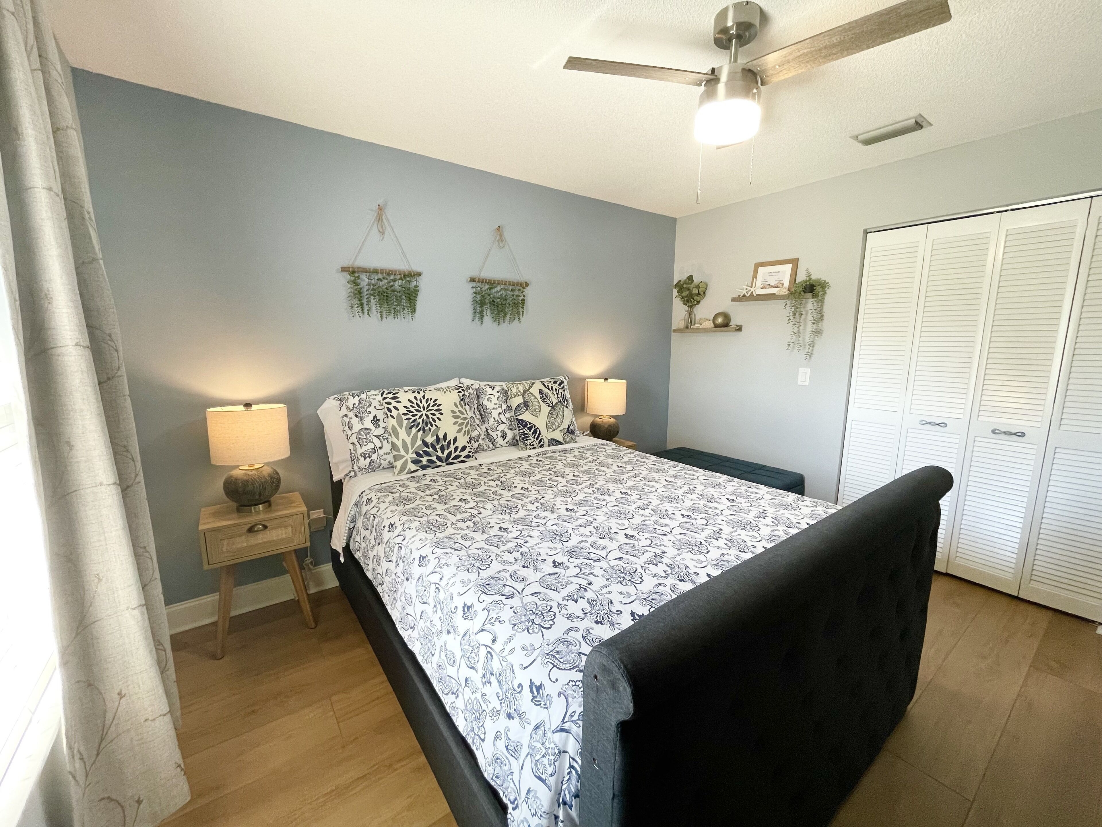 Quarto com cama, abajures, ventilador de teto, guarda-roupa e plantas decorativas.