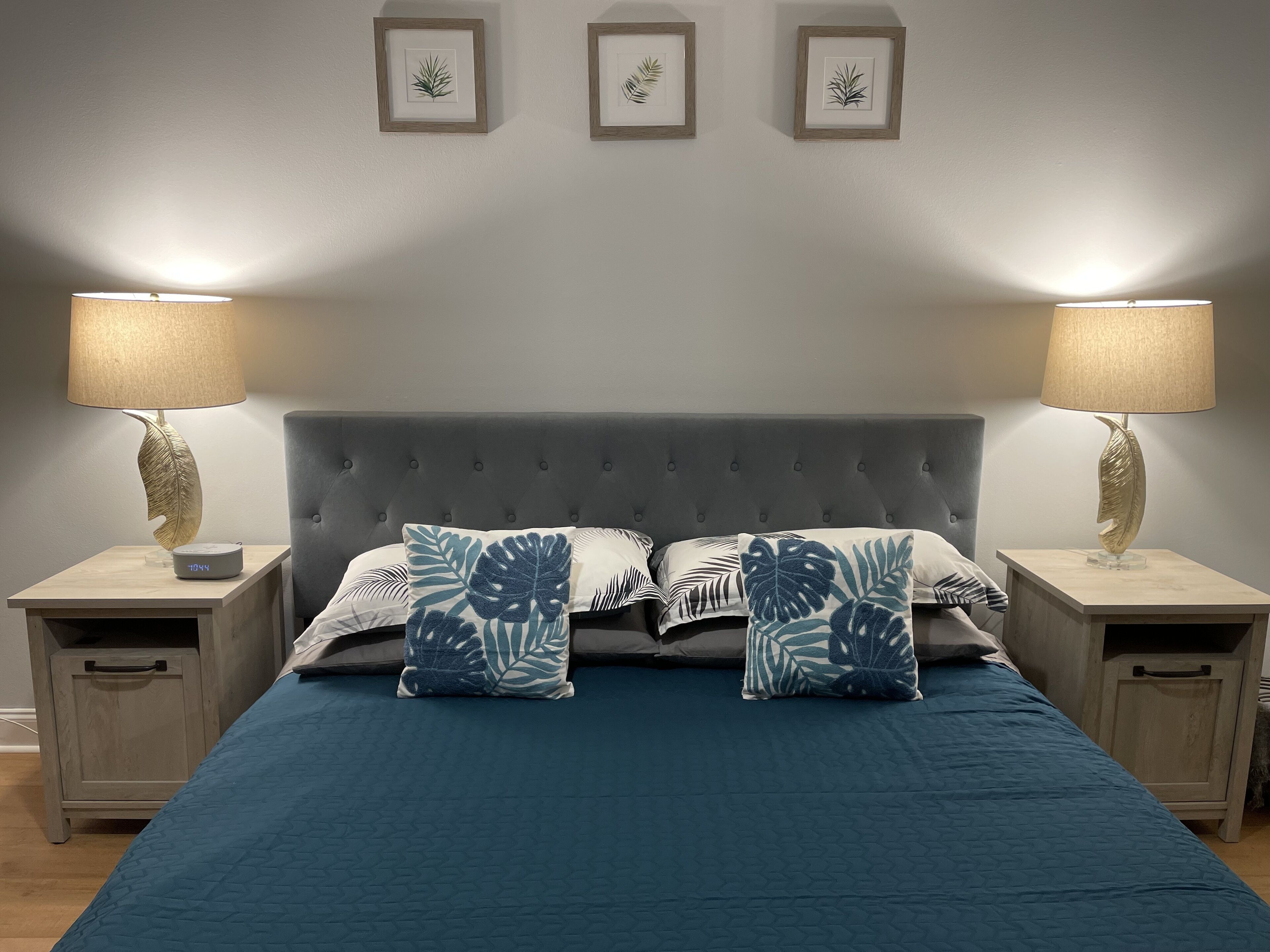 Un dormitorio con un cabecero gris capitonado, dos mesitas de noche con lámparas, y una cama con colcha azul y almohadas con estampado de hojas.