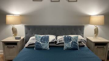Un dormitorio con un cabecero gris capitonado, dos mesitas de noche con lámparas, y una cama con colcha azul y almohadas con estampado de hojas.