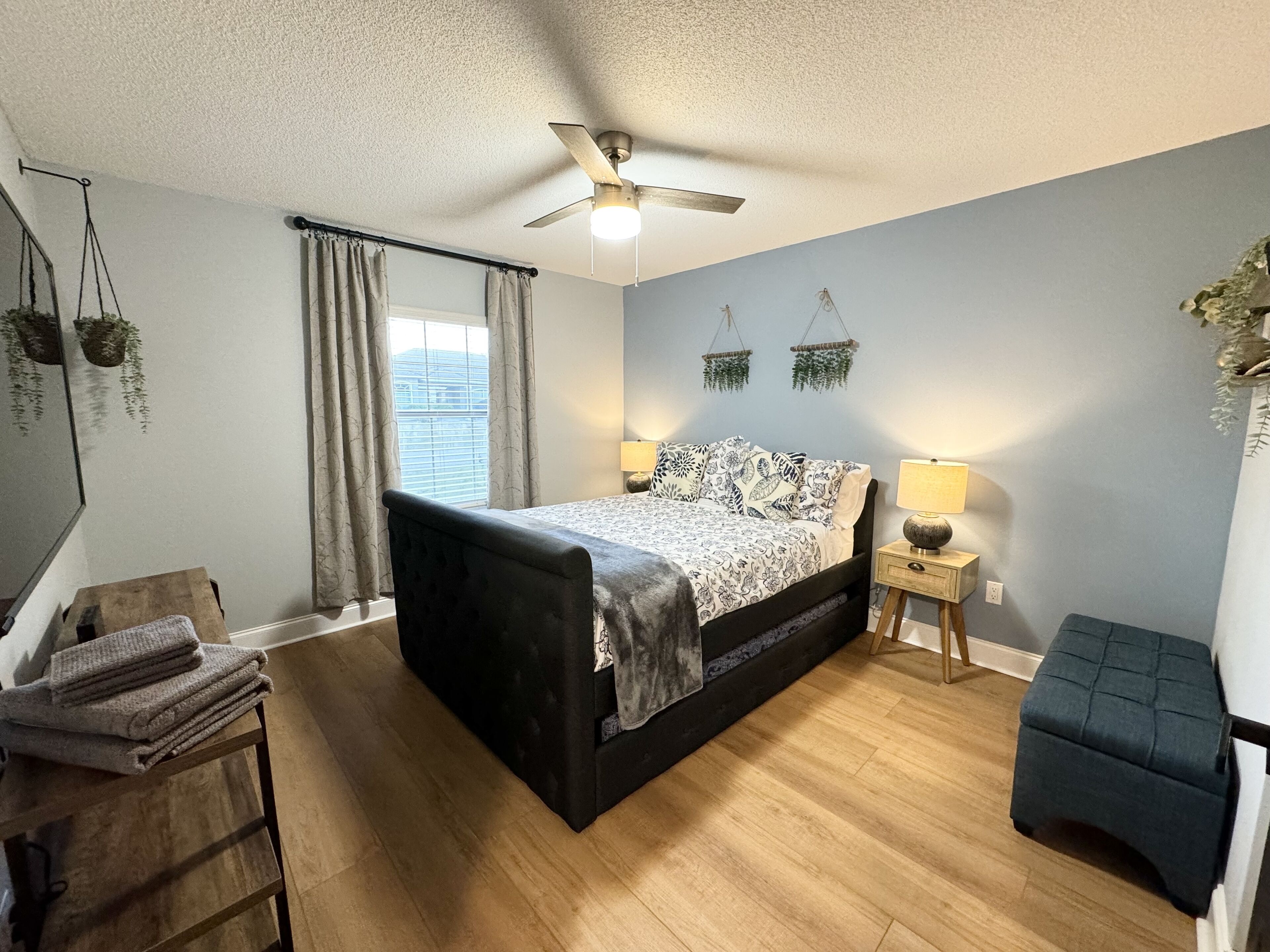 Une chambre à coucher comprenant un lit, des tables de chevet, un ventilateur de plafond, une fenêtre avec des rideaux et un petit tabouret.
