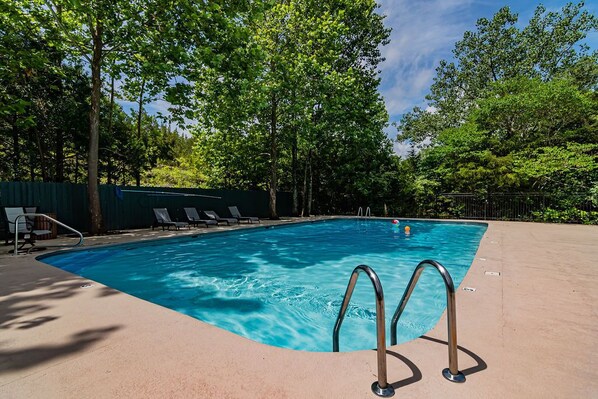 Une piscine rectangulaire à l’eau d’un bleu limpide, entourée d’une terrasse en béton. On y trouve des échelles métalliques du côté droit et une rangée de chaises longues vides le long du bord gauche.