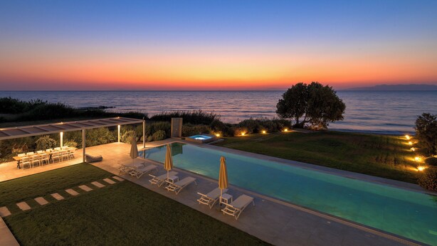 A pool area with lounge chairs and umbrellas, a covered seating area, and a view of the sea at sunset.