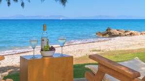 A wooden table with a bottle of wine, a glass, and a bowl of grapes on a beach with a view of the sea and distant mountains.