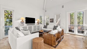A spacious living room with a white sofa, wicker furniture, a flat-screen TV, and large windows offering a view of the outdoors.