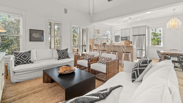 A spacious living area with a white sofa, a coffee table, and wicker chairs. A kitchen with modern appliances is visible in the background.
