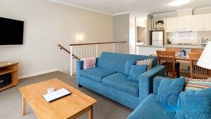 A living room with a blue sofa, a wooden coffee table, and a TV. There is a staircase with a railing and a kitchen area in the background. - Apollo Quays Apartment (Halls Head)