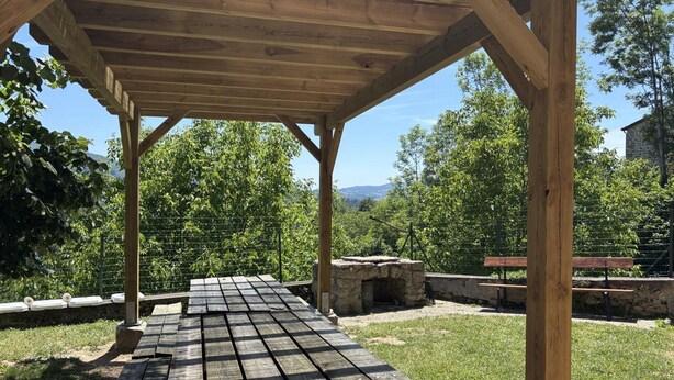 A wooden pergola with a picnic table underneath, surrounded by greenery and a stone wall.