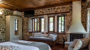 A rustic room with stone walls, a wooden ceiling, a bed with a patterned pillow, a bench with cushions, and a fireplace.