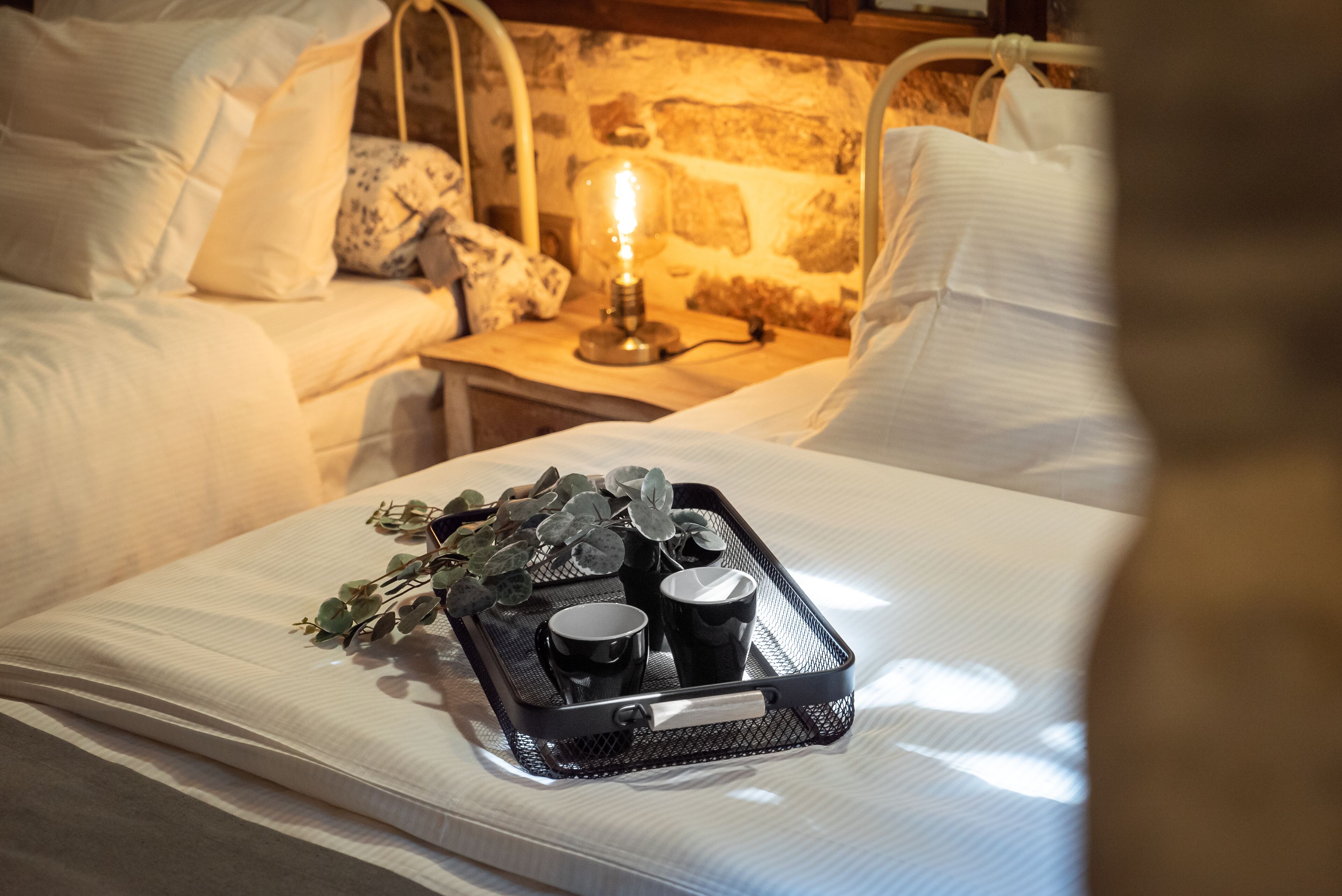 A neatly made bed with a tray of black cups and a candle on a bedside table.