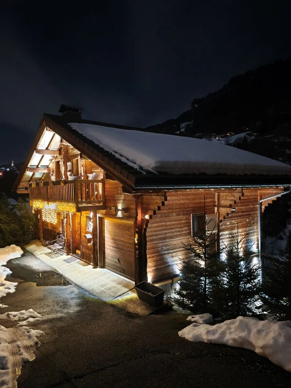 A wooden cabin with a sloped roof, illuminated windows, and a snow-covered ground.