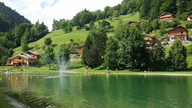 A lakeside view with wooden chalets, a fountain, and a grassy area with people. - La Chevrière, beautiful log chalet, panoramic view, near Megève (Flumet)