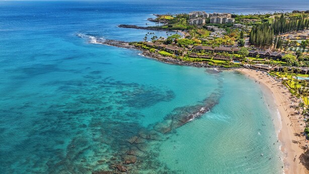 Vista aérea de un complejo turístico costero con aguas turquesas cristalinas, una playa de arena y un grupo denso de edificios con vistas al océano.