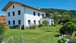 Une maison blanche à deux étages, dotée d’un balcon, entourée d’une pelouse et d’un jardin bien entretenus.