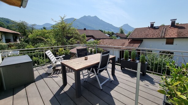 Une terrasse en bois avec une table et des chaises, entourée de plantes en pot et d’une rambarde en métal.