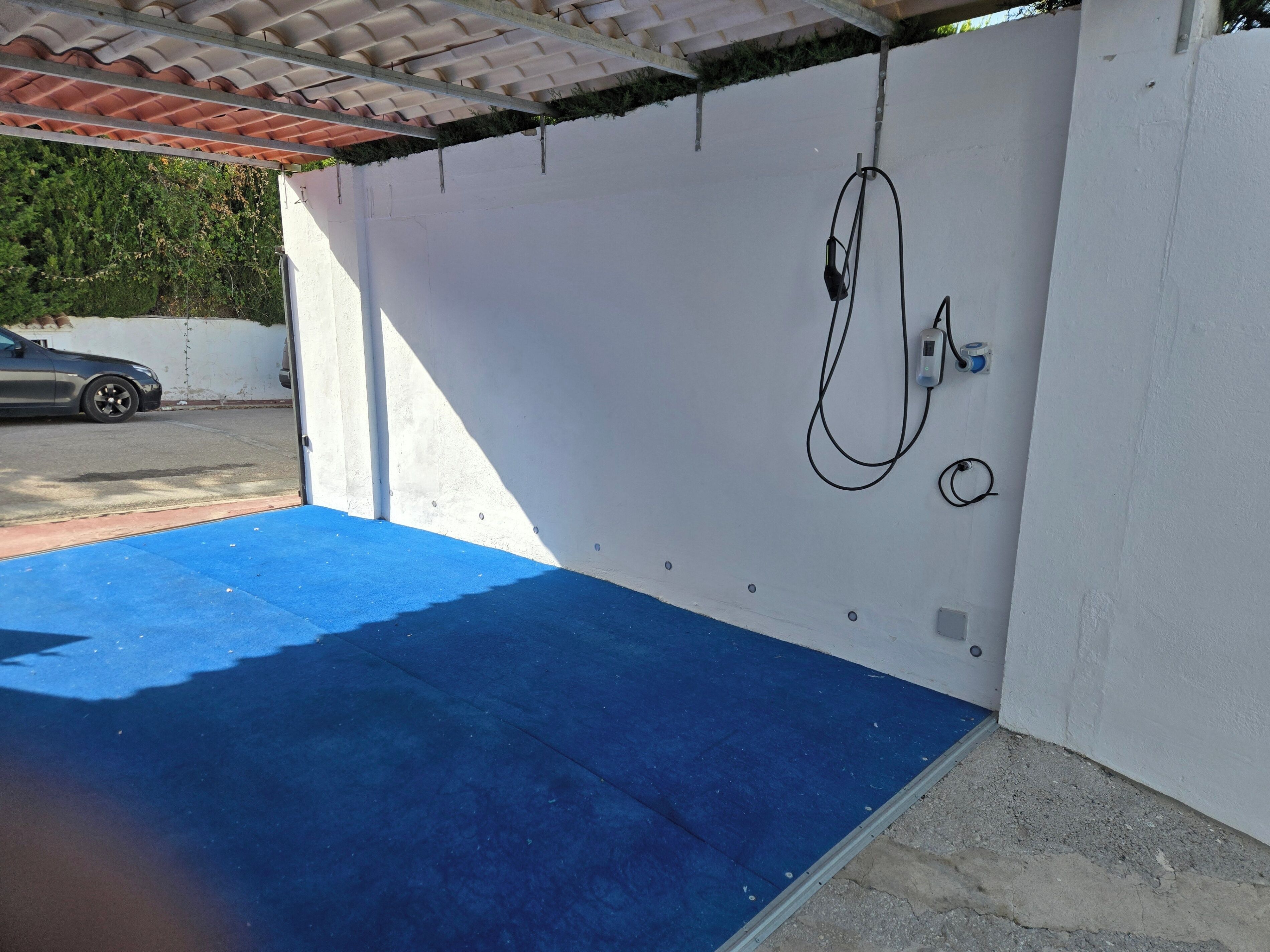 A covered outdoor area with a blue mat, a white wall, and a car parked outside.