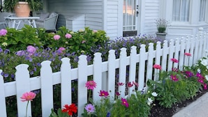 Une maison blanche, portant le numéro 107, avec une clôture en bois peinte en blanc et un jardin où s’épanouissent de nombreuses fleurs aux couleurs vives.