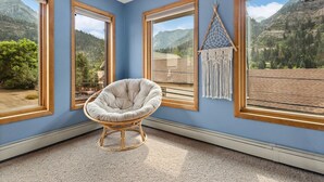 Ein Zimmer mit großen Fenstern und Blick auf die Berge, ein Rattan-Sessel und ein Wandbehang aus Makramee.