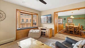 Ein Wohnzimmer mit Couch, Hocker und Fernseher an der Wand. Es gibt einen Essbereich mit Holztisch und Stühlen.