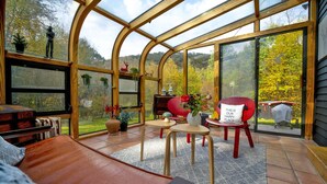 A conservatory with a curved wooden frame, large glass windows, and a view of a forested area. Inside, there is a wooden bench, a small table with a vase of flowers, and a red chair with a cushion.