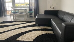 A living room with a black leather sofa, a black and white striped rug, a dining table with chairs, and a view of a body of water through large windows. - Vacation apartment Höhlenblick in Balve/Sauerland (Balve)