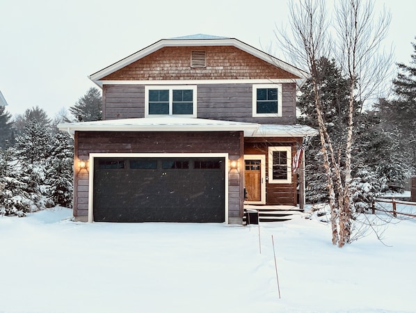 A two-story wooden house with a garage, surrounded by snow-covered trees.