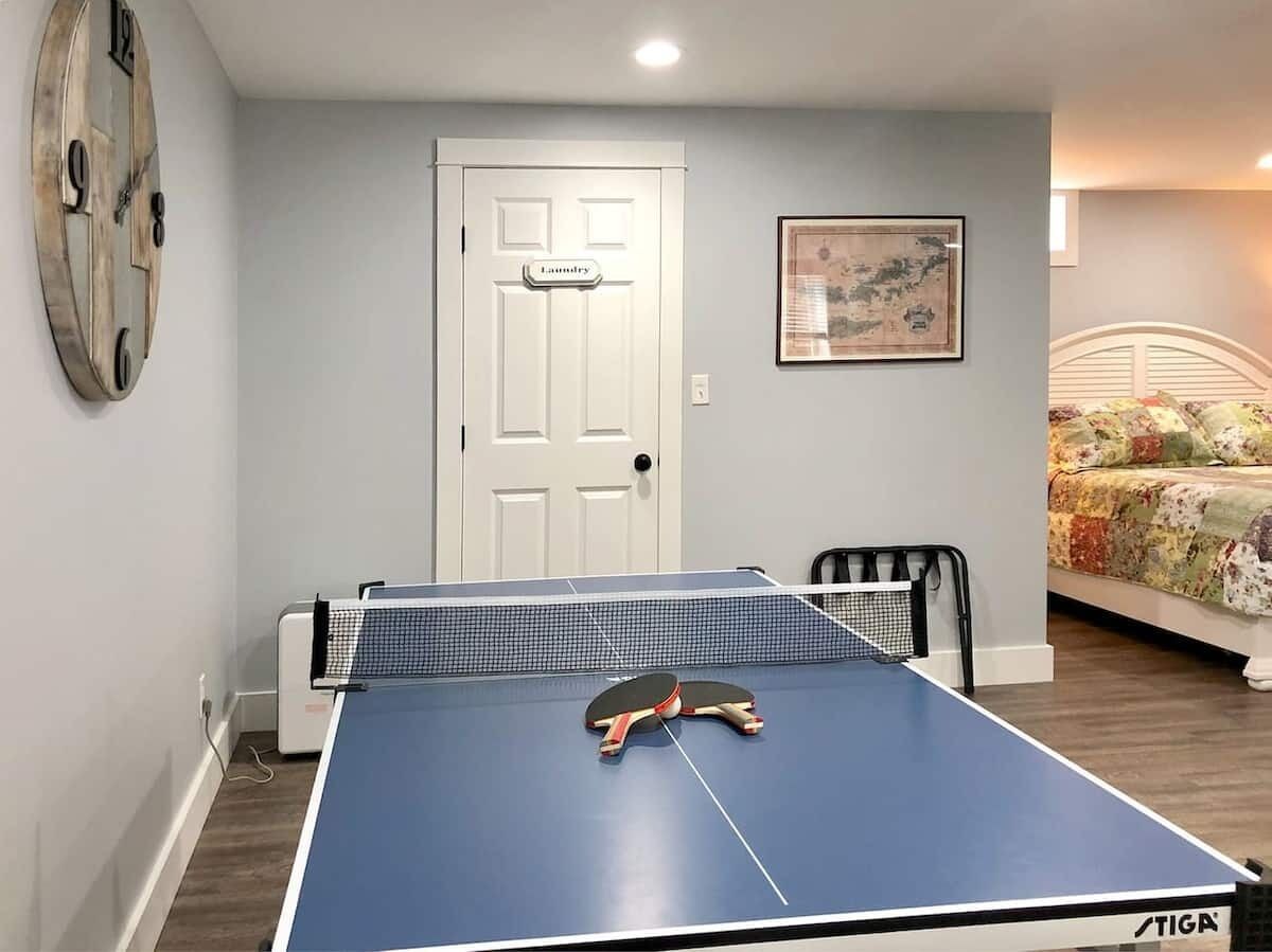 Une pièce avec une table de ping-pong, une porte portant l’inscription « Buanderie », un tableau encadré au mur et un lit recouvert d’une courtepointe à motifs floraux.