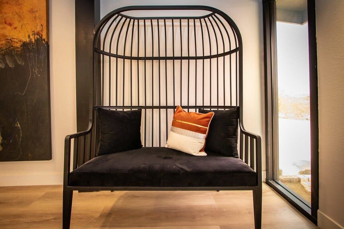A modern chair with a black metal frame and dark cushions, positioned in a room with a large window and a piece of abstract art on the wall.