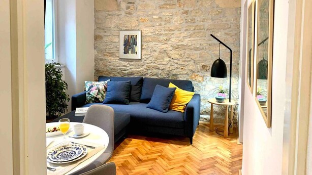 A cozy living room with a stone wall, a blue sofa with yellow cushions, a round table with a white plate and a glass of orange juice, and a potted plant. - City Center Appartment Near Arena-pula (Pula)